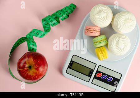 Red Apple mit grünen Maßband, digitale Küchenwaage mit Macarons und Baisers auf rosa Hintergrund. Gewicht Verlust und schlankheitsbehandlung Konzept Stockfoto