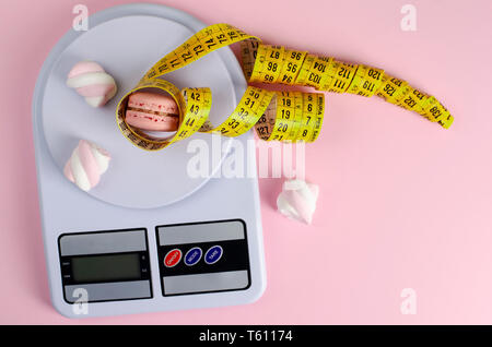 Gelbe Maßband, digitale Küchenwaage mit Macarons und Marshmallows auf Pastell rosa Hintergrund. Gewichtsabnahme und schlankheits Behandlungskonzept. Zu Stockfoto