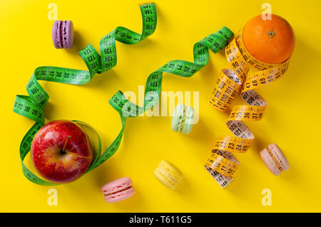 Roter Apfel und Orange mit Maßbändern und süßen französischen Makronen auf gelben Hintergrund. Gewichtsabnahme und schlankheits Behandlungskonzept. Keine Diät Tag Stockfoto