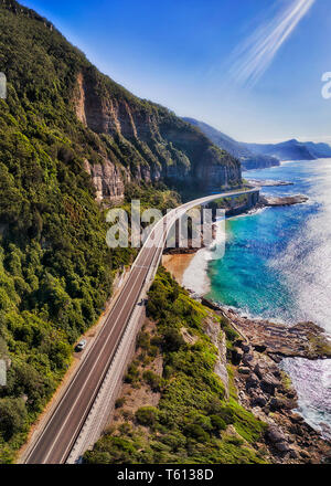 Grand Pacific Drive Segment der Sea Cliff Brücke um steile Sandsteinfelsen über Wellen des Pazifischen Ozeans an einem sonnigen Tag in vertikale Panorama. Stockfoto