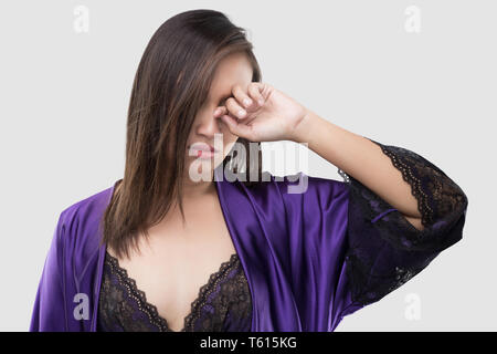Die Frau im Nachthemd Seide und Spitze Gewand reibt ihr Auge mit einem Finger gegen den grauen Hintergrund, Frauen in Violett satin Nachtwäsche, Juckreiz Stockfoto
