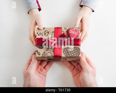 Die Hände des Kindes und die Hände des erwachsenen Mannes, schönen Geschenkkarton, Farbband und glasierte Plätzchen auf einem weißen, Holz- Hintergrund. Ansicht von oben, close-up. Vorbereitung für Th Stockfoto