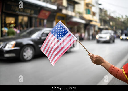 Woman's Hand hält die USA-Flagge auf dem verschwommenen Hintergrund des fahrenden Autos der amerikanischen diplomatischen Escort auf der Straße der Stadt während des Politischen gegenüber Stockfoto