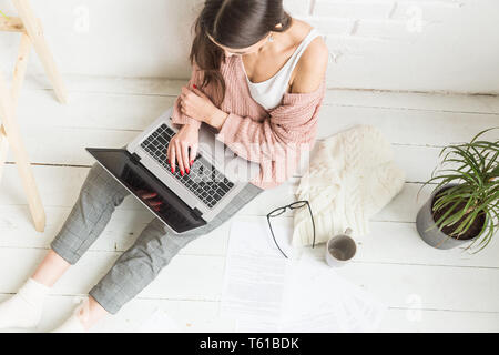 Eine junge Frau sitzt auf dem Boden in einem hellen Wohnung oder Büro innen und arbeitet an einem Laptop, Freelancer Mädchen bei der Arbeit, das Konzept der Abstand Stockfoto