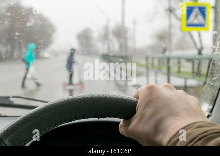 Nahaufnahme, Hand des Fahrers auf das Lenkrad des Autos auf dem Hintergrund einer Fußgängerampel im regnerischen Wetter und eine Mutter mit einem Kind auf Stockfoto
