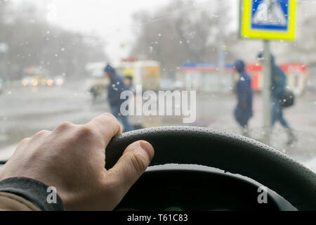 Blick aus dem Auto, Hand des Mannes am Lenkrad des Autos, befindet sich gegenüber der Fußgängerampel und Fußgänger über die Straße Stockfoto