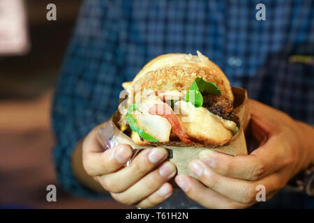 Hand des Menschen nehmen grossen Hamburger, Speck, halten und bereit sind, ihn in seinen Mund zu beißen, bei Thailand foodtruck Ereignis. Stockfoto