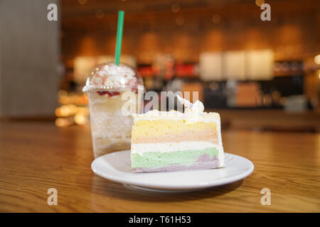 Rutsche rainbow Kuchen dekorieren mit Unicorn Horn auf der Oberseite neben Kaffee frappe. Der Kuchen ist in eine weiße Scheibe auf dem Holztisch und Unschärfe im Hintergrund. Stockfoto