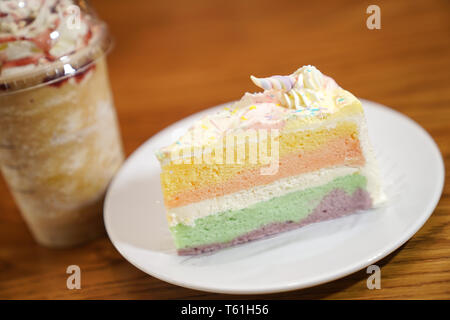 Rutsche rainbow Kuchen dekorieren mit Unicorn Horn auf der Oberseite neben Kaffee frappe. Der Kuchen ist in eine weiße Scheibe auf dem Holztisch und Unschärfe im Hintergrund. Stockfoto
