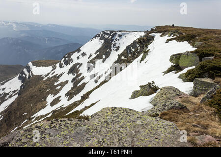 Vordergrund Rock von grünen und gelben Moos und Rocky Mountain Cliff teilweise durch Schnee während der Übergang zwischen Winter und Frühling fallen Stockfoto