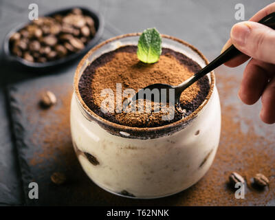 Hohe Betrachtungswinkel von tiramisu in Glas Schale auf Steinplatte über schwarzen konkreten Hintergrund, Hand mit Teelöffel isst Tiramisu. Kopieren Sie Platz. Stockfoto