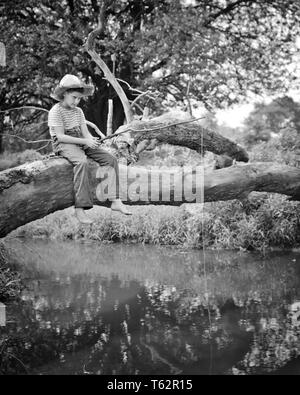 1940er barfuß Land Junge trägt blaue Jeans gestreiftes Hemd und STROHHUT sitzend auf einen umgestürzten Baum ÜBER EINEN BACH ANGELN - ein 64 HAR 001 HARS SERENITY B&W SKILL AKTIVITÄT UNTERHALTUNG ABENTEUER GEFALLEN HOBBY FREIZEIT UND HOBBYS WISSEN FREIZEIT ZEITVERTREIB VERGNÜGEN AUF BARFUSS STROHHUT HUCK FINN T-SHIRT BLUE JEANS TOM SAWYER HUCKLEBERRY FINN JUGENDLICHE ENTSPANNUNG AMATEUR SCHWARZ UND WEISS KAUKASISCHEN ETHNIE CREEK GENUSS HAR 001 ALTMODISCH Stockfoto