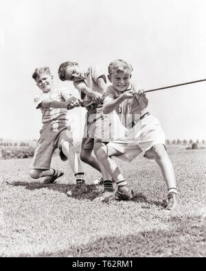 1930er Jahre 1940er Jahre drei lächelnd energetische jugendlich Jungen kämpfen zusammen zieht das Seil im TAUZIEHEN TRAGEN SOMMER Shorts und Turnschuhen - b4409 HAR 001 HARS LÄNDLICHEN GESUNDHEIT LEICHTATHLETIK KOPIE RAUM FREUNDSCHAFT IN VOLLER LÄNGE RISIKO ATHLETISCHE SNEAKERS VERTRAUEN B&W SOMMER AKTIVITÄT GLÜCK KÖRPERLICHE KRAFT SIEG MUT UND WETTBEWERB ANSTRENGUNG AUFREGUNG KRÄFTIGE ERHOLUNG IN AUF JUGENDLICHE KONZEPTIONELLE ATHLETEN FLEXIBILITÄT MUSKELN STILVOLLE KURZE HOSEN ZUSAMMENARBEIT ENERGISCHER PRE-TEEN PRE-TEEN BOY KÄMPFEN MITEINANDER TAUZIEHEN SCHWARZ UND WEISS KAUKASISCHEN ETHNIE HAR 001 ALTMODISCH Stockfoto