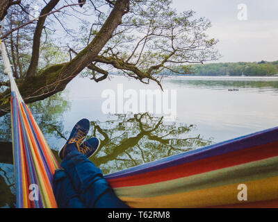 Mann mit blauen Schuhe und Jeans schwingt in farbenfrohen Hängematte hängt am Ufer des Sees in Dänemark Stockfoto