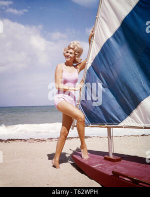 1960 s Frau mittleren Alters mit BLONDEN WIND GROSSE HAAR ZU TRAGEN ROSA ONE PIECE BADEANZUG STÄNDIGEN von Sail yacht am Ocean Beach - kg 2694 SCH002 HARS SEGEL FRÖHLICH DURCH AUF LÄCHELN KONZEPTIONELLE GEBLASEN FREUDIGE SEGELSCHIFF MITTELALTER BADEANZUG MITTE NACH MITTE DER ERWACHSENEN FRAU GROSSEN HAAR KAUKASISCHEN ETHNIE ALTMODISCH EIN STÜCK WIND AUSGEBLASEN Stockfoto