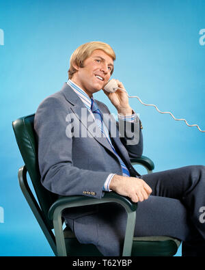 1970 s BLONDE GESCHÄFTSMANN IM BÜRO STUHL GESPRÄCH AM TELEFON MIT KAMERA-ks 8210 HAR 001 HARS GERNE FREUDE LIFESTYLE ZUFRIEDENHEIT STUDIO SHOT KOPIE RAUM IN VOLLER LÄNGE mit halber Länge PERSONEN MÄNNER VERTRAUEN AUSDRÜCKE AUGENKONTAKT ERFOLG Anzug und Krawatte verkaufen, fröhlichen STRATEGIE CUSTOMER SERVICE NETWORKING FÜHRUNG FORTSCHRITTE STOLZ AUF AUTORITÄT BERUFEN LÄCHELT VERBINDUNG KONZEPTIONELLER FREUDIGE stilvolle Mitte - Mitte - erwachsenen Mann VERKÄUFER KAUKASISCHEN ETHNIE HAR 001 ALTMODISCH Stockfoto