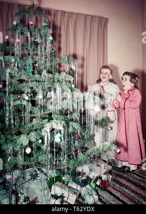 1950er Jahre zwei Mädchen VOR DEM CHRISTBAUM MIT BLICK DER WUNDER TRAGEN SCHLAFANZUG - kx971 FST001 HARS FAMILIEN FREUDE LIFESTYLE FRAUEN GESUNDHEIT LEBEN ZU HAUSE KOPIEREN RAUM VOLLER LÄNGE INSPIRATION SCHLAFANZUG GESCHWISTER EINGERICHTET SCHWESTERN AUSDRÜCKE WUNDER GLÜCK ENTDECKUNG FROHE AUFREGUNG GESCHWISTER LÄCHELN DEZEMBER KONZEPTIONELLE 25. Dezember fröhliche FRÖHLICHE JUGENDLICHE MITEINANDER KAUKASISCHEN ETHNIE ALTMODISCH Stockfoto