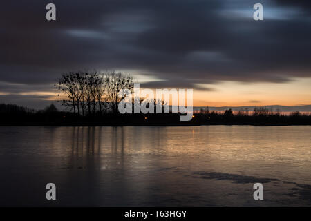 Bewölkt dunkle Wolken nach Sonnenuntergang auf einem zugefrorenen See und springenden Bäumen im Eis - Abendlicher Blick Stockfoto