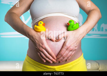 Junge schwangere Frau, ihren Bauch zu Hause zeigen schön abgerundet, während sie mit den Händen schöne Herz und Spielen mit bunten Ente Spielzeug während Stockfoto