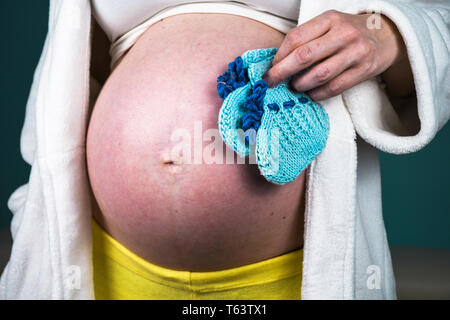 Junge schwangere Frau, ihren Bauch zu Hause zeigen schön abgerundet, während Sie in der Hand kleine blaue gestrickte Baby Socken Stockfoto