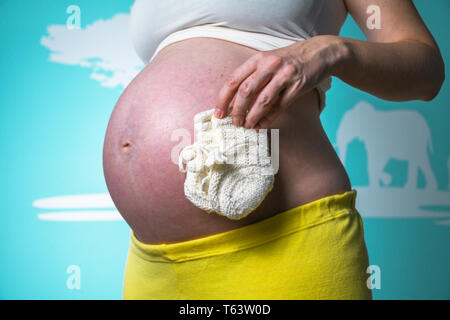 Junge schwangere Frau, ihren Bauch zu Hause zeigen schön abgerundet, während Sie in der Hand kleine weiße gestrickte Baby Socken Stockfoto