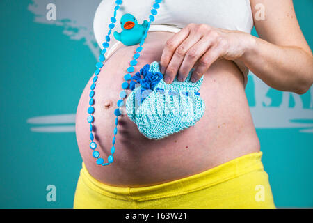 Junge schwangere Frau, ihren Bauch zu Hause zeigen schön abgerundet, indem die Hand blau Halskette, einem Kunststoff ente Spielzeug und gestrickte kleine Baby Schuhe Stockfoto