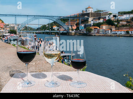 Selektiver Fokus der Roten, Weißen und Porto Weingläser mit Blick auf Cais da Ribeira und Ponte de Dom Luis I auf dem Douro in Porto, Portugal Stockfoto