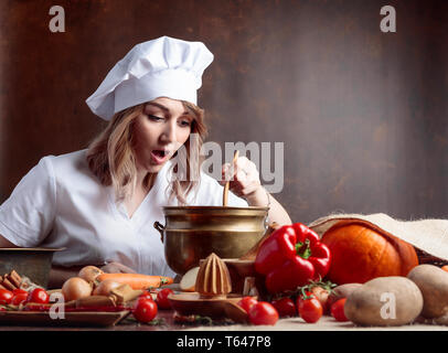 Junge Frau in einem Küchenchef Uniform mit altes Messing Topf und Löffel aus Holz. Auf einem Tisch verschiedenen Küchenutensilien und Gemüse. Stockfoto