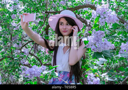 Mädchen nimmt Bilder von sich selbst am Telefon, Kommunikation in sozialen Netzwerken, eine Videokonferenz, sitzend auf einem Zweig der blühenden Flieder Stockfoto