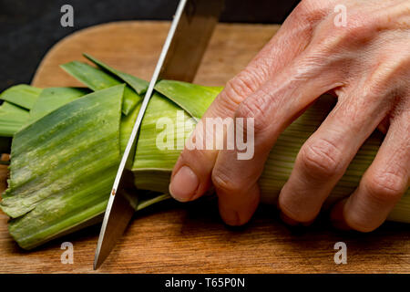 Nahaufnahme von einem Koch bereitet frische organische Lauch (Allium ampeloprasum) mit einem scharfen Messer auf einer hölzernen Schneidebrett. Stockfoto