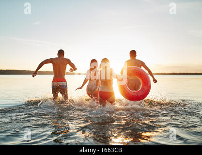 Rückspiegel der Gruppe von verschiedenen jungen Freunde in Badebekleidung in einen See zusammen laufen bei Sonnenuntergang plantschen und Wellen Stockfoto