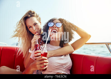 Zwei lachende Junge weibliche Freunde Drinks, während auf einem Boot sitzen zusammen auf dem offenen Meer in den Sommerferien Stockfoto