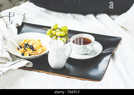 Frühstück im Bett. Cornflakes mit Beeren und Milch, Früchte und Tee auf einem schwarzen Fach. Stockfoto