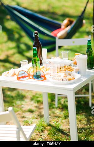 Tabelle mit Flaschen Bier und Essen auf Sommer Garden Party - die Frau, die in einer Hängematte im Hintergrund Stockfoto