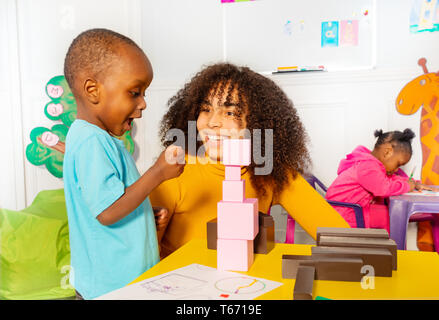 Aufgeregten kleinen schwarzen Jungen im Kindergarten über Turm von Blöcken zusammen mit Lehrer Stockfoto