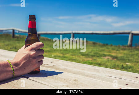 Ferienhäuser Konzept. Männliche Hand, die Flasche Bier auf dem Meer Strand auf der hölzernen Plattform Stockfoto