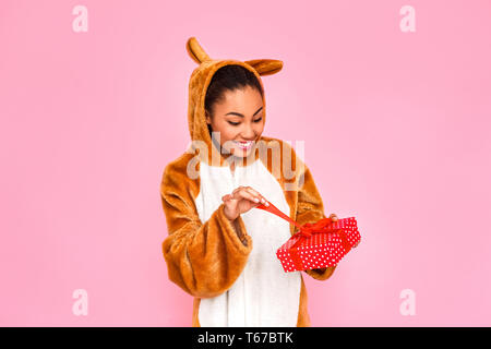 Junge Frau in Bunny kigurumi Ständigen isoliert auf rosa Hintergrund holding Ribbon öffnung Rot vorhanden, lächelt überrascht Stockfoto