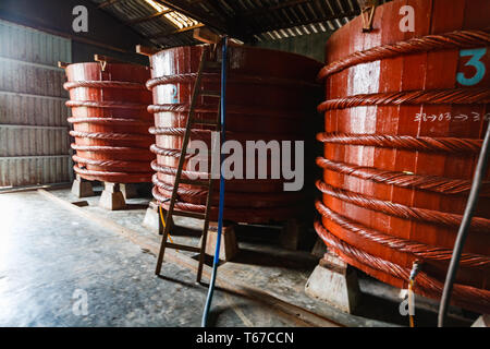 Drei große, rote Holzfässer, verstärkt mit dickem Seil, stehen in einer Reihe in einem Gebäude, wahrscheinlich in einer Fabrik oder einem Lager, mit einer Leiter, die gegen den mittleren behälter anliegt und möglicherweise für Wartung oder Zugang verwendet wird. Stockfoto