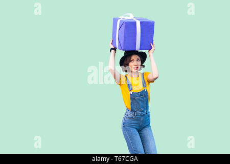 Träumen positive junges Mädchen in hipster Verschleiß in Jeans Overalls und schwarzen Hut und Holding unter dem Kopf riesen große schwere Geschenkbox mit dem Erwägen f Stockfoto