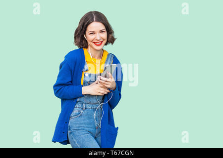 Porträt der Schönen fröhlicher junger erwachsenen Frau in hipster Verschleiß in Jeans Overalls, die hielten Telefon und hören Musik mit Mobilteilen Stockfoto