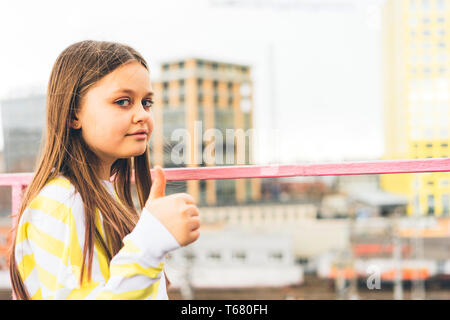 Ein junges Mädchen in einem gelben Pullover steht vor dem Hintergrund des Stadtbildes und zeigt eine Hand mit einem angehobenen Finger Stockfoto