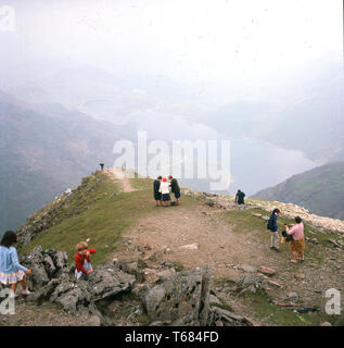 1960, historische, Erwachsene und Kinder auf dem Gipfel des Mount Snowdon, der höchste Berg in Wales. Sie sind alle normalen, alltäglichen Kleidung tragen, als wenn für einen Spaziergang im Park und nicht Spezialist Outdoor Gear, würde man erwarten, dass Sie heute zu tragen. Da der Snowdon ist der höchste Berg in Großbritannien (ohne Ben Nevis) und das Wetter könnte jeden Moment ändern, es zeigt, wie die Leute besucht wurden und Sehenswürdigkeiten Sehenswürdigkeiten in diesem Jahrzehnt später auf. Stockfoto