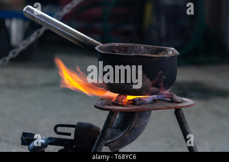 Close-up Metallwanne ist beheizt auf einem Gasbrenner in einem Metall schmelzen Workshop von führen. Travel Option kochen in einer Pfanne auf einem Gasbrenner. Eisen und Stahl Stockfoto