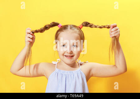 Closeup Portrait von ein fröhliches kleines Mädchen auf einem gelben Hintergrund. Das Kind sieht in die Kamera und hält seine Hände Pigtails aus Haar. Stockfoto