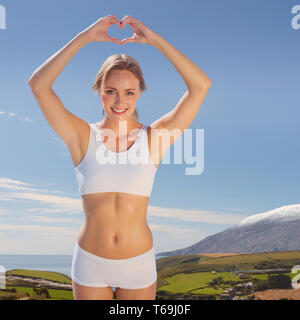 Zusammengesetztes Bild des wunderschönen passen blonde die Herzform mit Händen am Meer Stockfoto