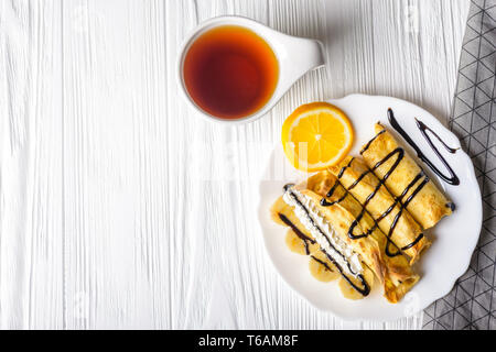 Pfannkuchen mit Banane, Sahne und Schokolade Sirup auf weißem Holz- Hintergrund und die Tasse Kaffee. Stockfoto