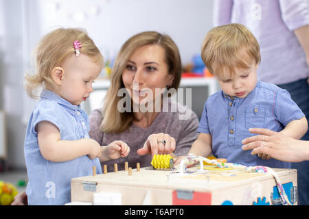 Cute Kleinkinder Babys spielen mit busyboard. Mutter oder Betreuer Unterricht für Kinder im Kindergarten. Pädagogisches Spielzeug für Kinder. Stockfoto