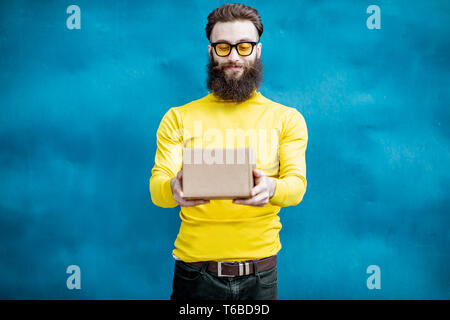 Stilvolle bärtigen Mann in gelb Pullover mit Geschenkbox auf dem blauen Hintergrund Stockfoto