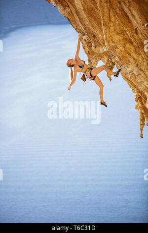 Junge weibliche Kletterer im Bikini hängen mit der einen Hand unter überhängenden Felsen. Auf anspruchsvolle Kletterroute über Meer. Stockfoto