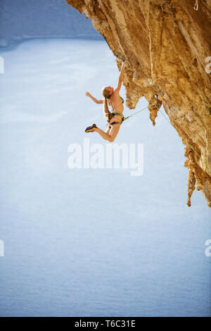 Junge weibliche Kletterer im Bikini hängen mit der einen Hand unter überhängenden Felsen. Auf anspruchsvolle Kletterroute über Meer. Stockfoto